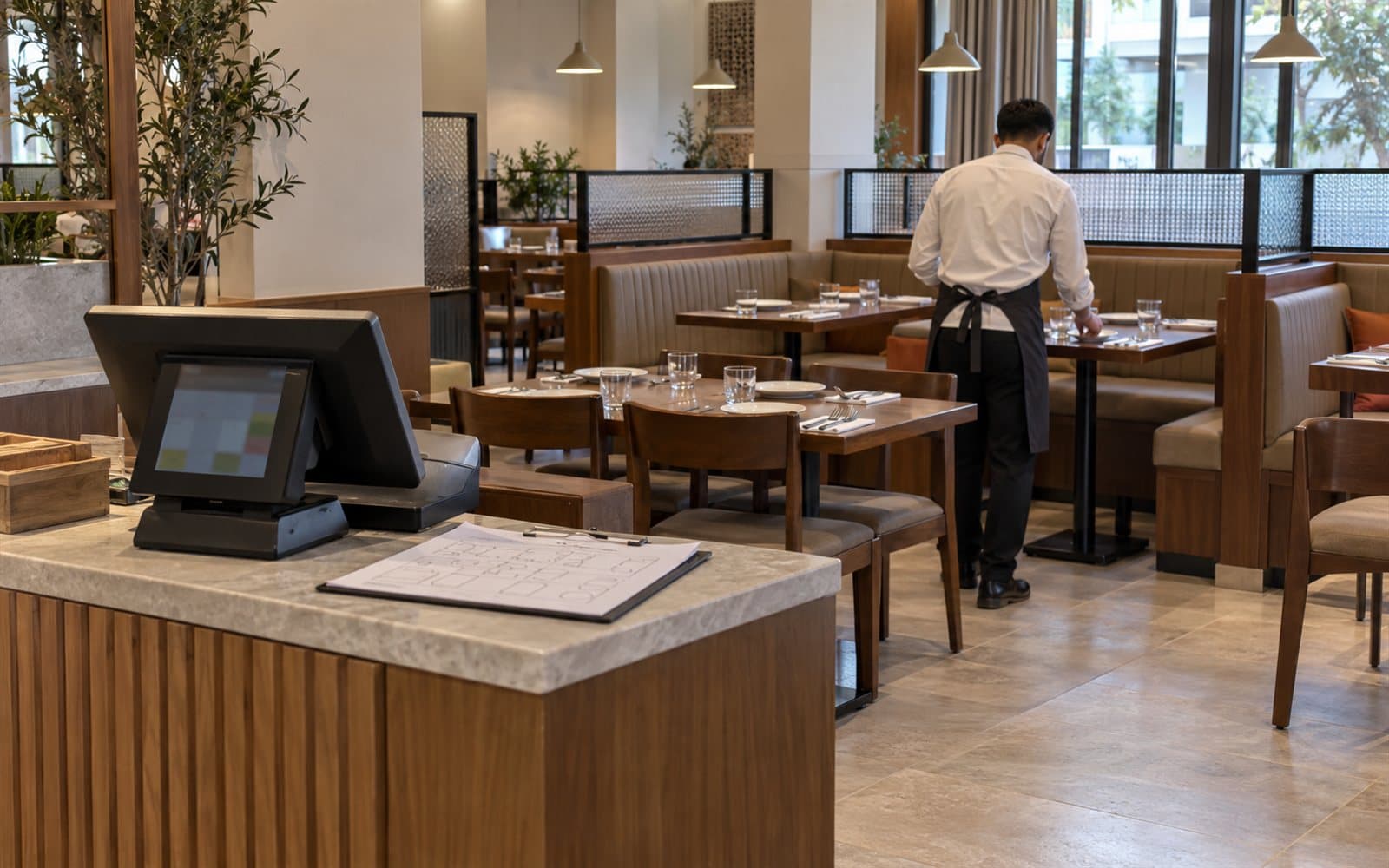 Restaurant dining room before service, with the host stand POS in front and a waiter setting tables
